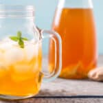 A mason jar mug filled with ice and a ginger switchel drink, garnished with a sprig of spearmint. There is a carafe of switchel and some ginger and spearmint in the background.