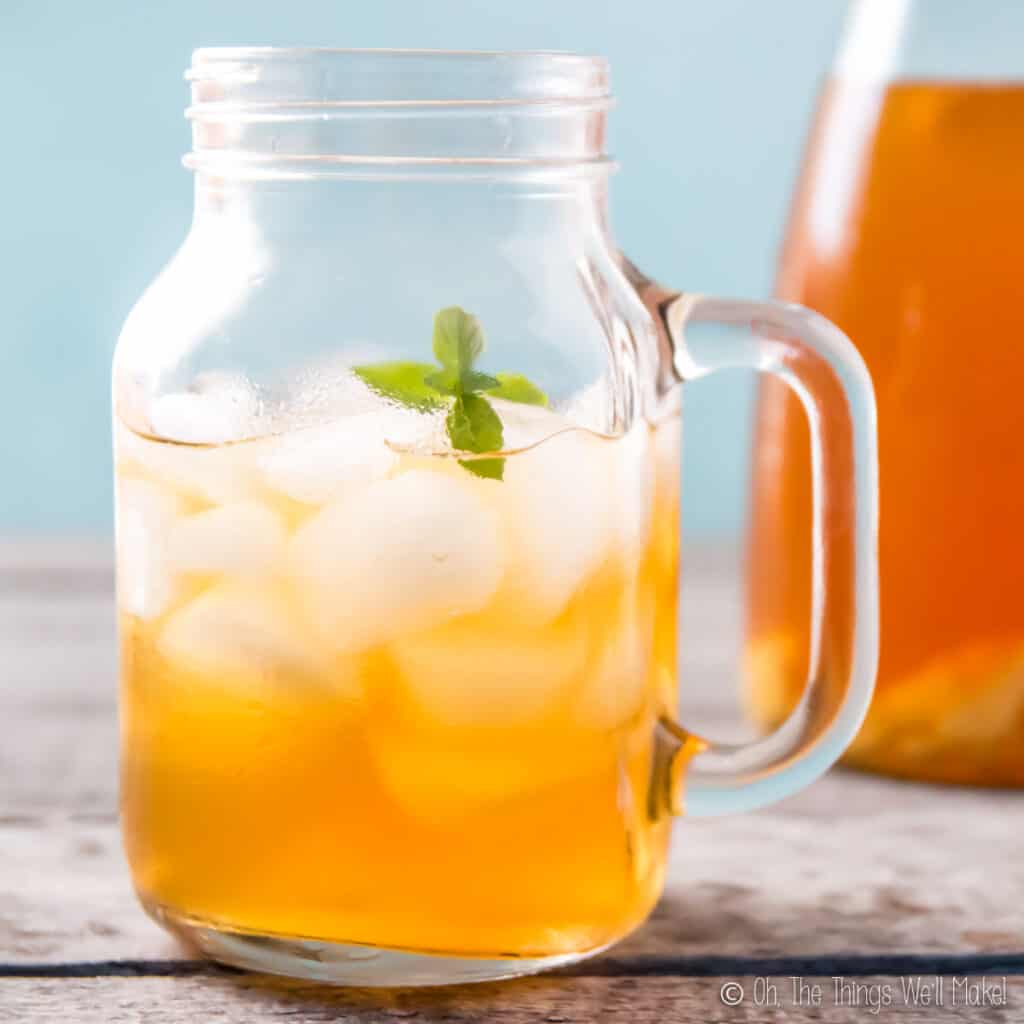 A mason jar mug filled with ice and a ginger switchel drink, garnished with a sprig of spearmint. There is a carafe of switchel in the background.