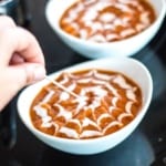 Running a toothpick from the center to the outer concentric circles of greek yogurt on a bowl of roasted pumpkin soup to form a spiderweb design.