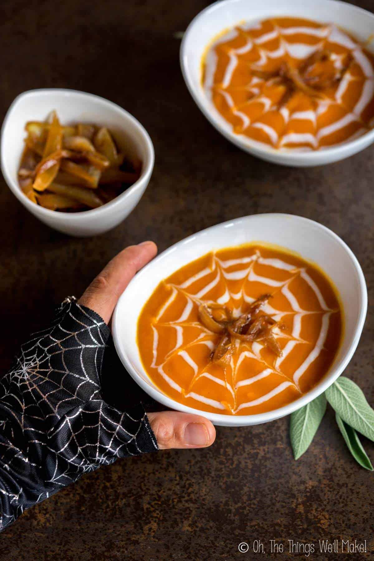 A gloved hand reaching for a roasted pumpkin soup decorated for Halloween with a cream spiderweb and caramelized onion "spiders." 