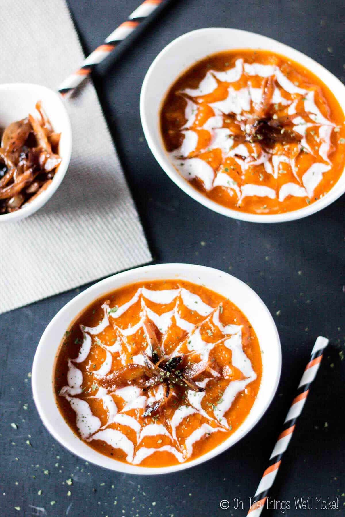 Overhead view of two bowls of a roasted pumpkin and red pepper soup decorated with green yogurt in a spiderweb design. They're garnished with caramelized onions, made to look like spiders on the web..