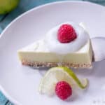 Overhead view of a slice of a paleo key lime pie garnished with coconut cream and a raspberry being placed on a white plate with a silver spatula next to a slice of lime and a raspberry.