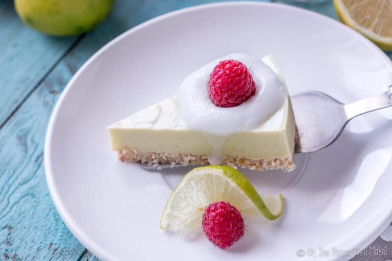 Overhead view of a slice of a paleo key lime pie garnished with coconut cream and a raspberry being placed on a white plate with a silver spatula next to a slice of lime and a raspberry.