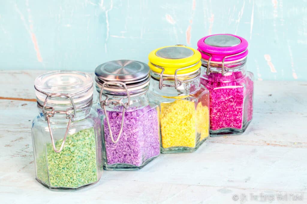 4 glass jars filled with 4 different types of colorful sprinkles made from shredded coconut. From left to right: green, purple, yellow, and bright pink.