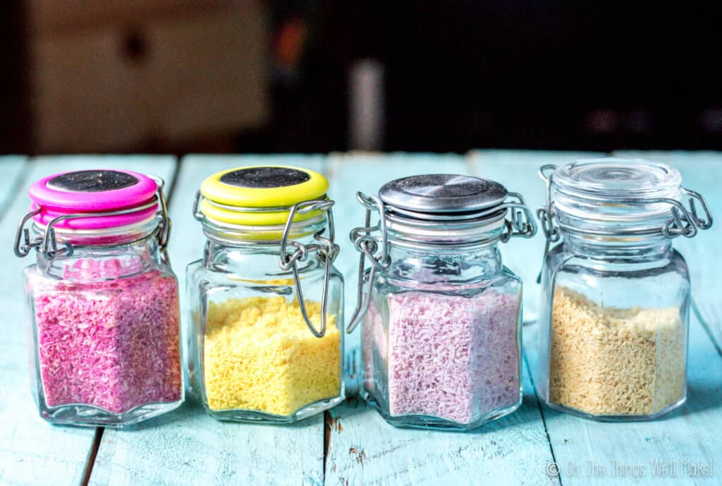 4 glass jars filled with 4 different types of colorful sprinkles made from shredded coconut after stored for 10 months. The colors have faded, but are still visible. From left to right: bright pink, yellow, purple, and what was once green, but is now more beige.