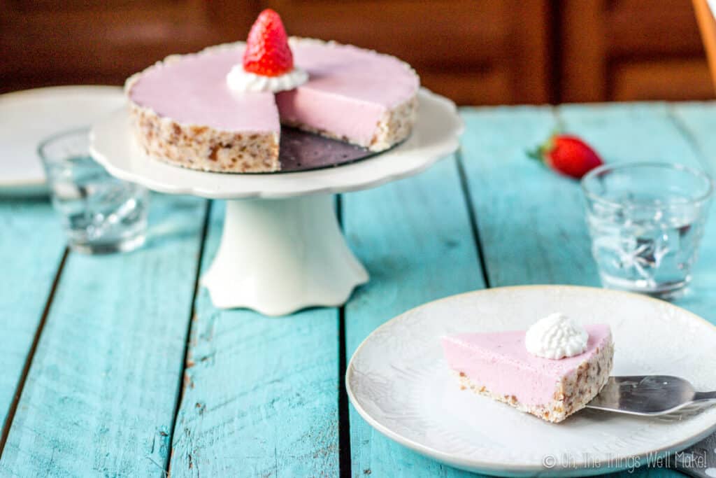 A paleo strawberrry cream pie on a cake stand with a slice missing. The slice is being places on a light beige plate with a spatula.