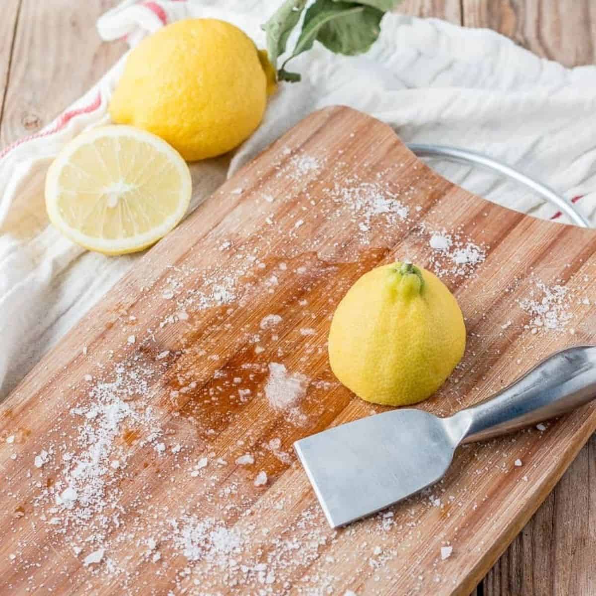Sliced lemons and stainless steel scraper on a wooden chopping board with some baking soda scattered around it.