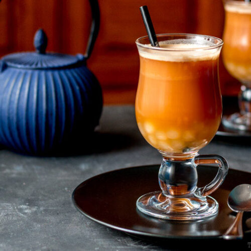 Several chai lattes with homemade boba in front of a blue iron teapot.