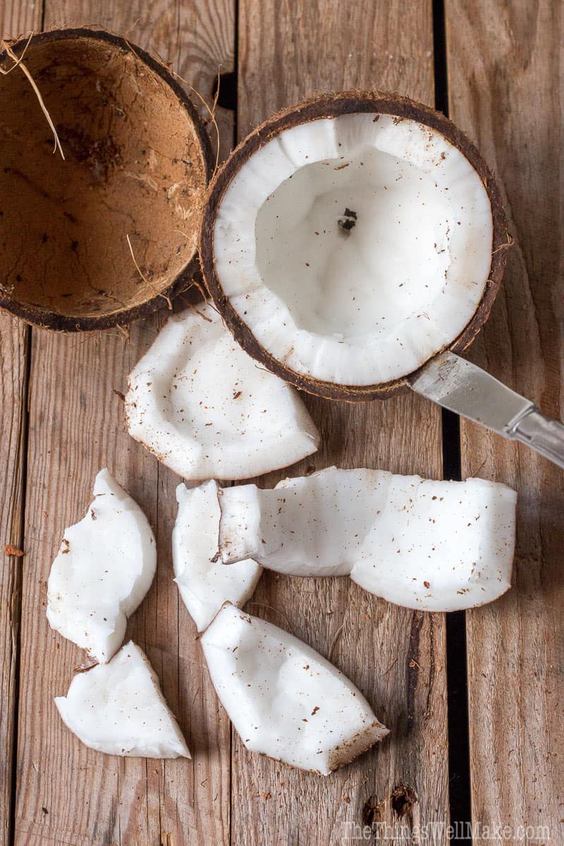 Ovehead view of an empty coconut shell and a knife running between the shell and meat of a coconut half, next to various pieces of coconut