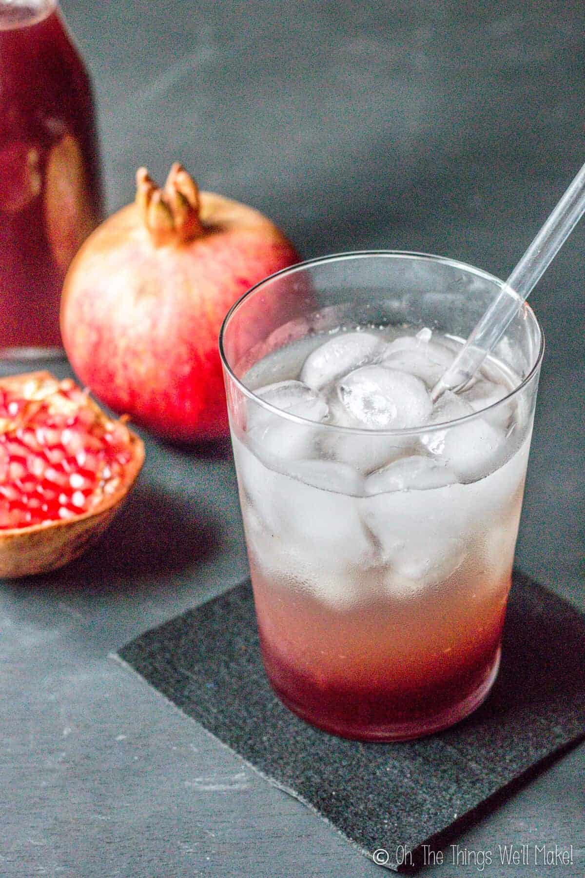 Closeup of a glass of soda and grenadine over ice in front of some pomegranates and a bottle of homemade grenadine.