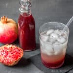 A closeup of a glass of soda and grenadine over ice in front of pomegranates and a bottle of homemade grenadine.