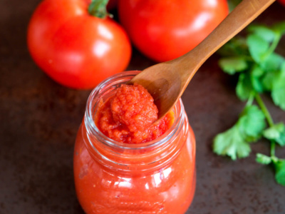 Over head view of a spoon full of tomato paste over a jar full of it. A couple of fresh tomatoes lay next to the jar.