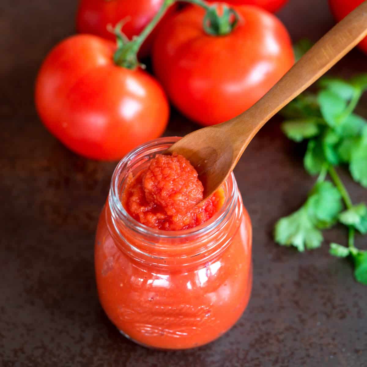 Over head view of a spoon full of tomato paste over a jar full of it. A couple of fresh tomatoes lay next to the jar.