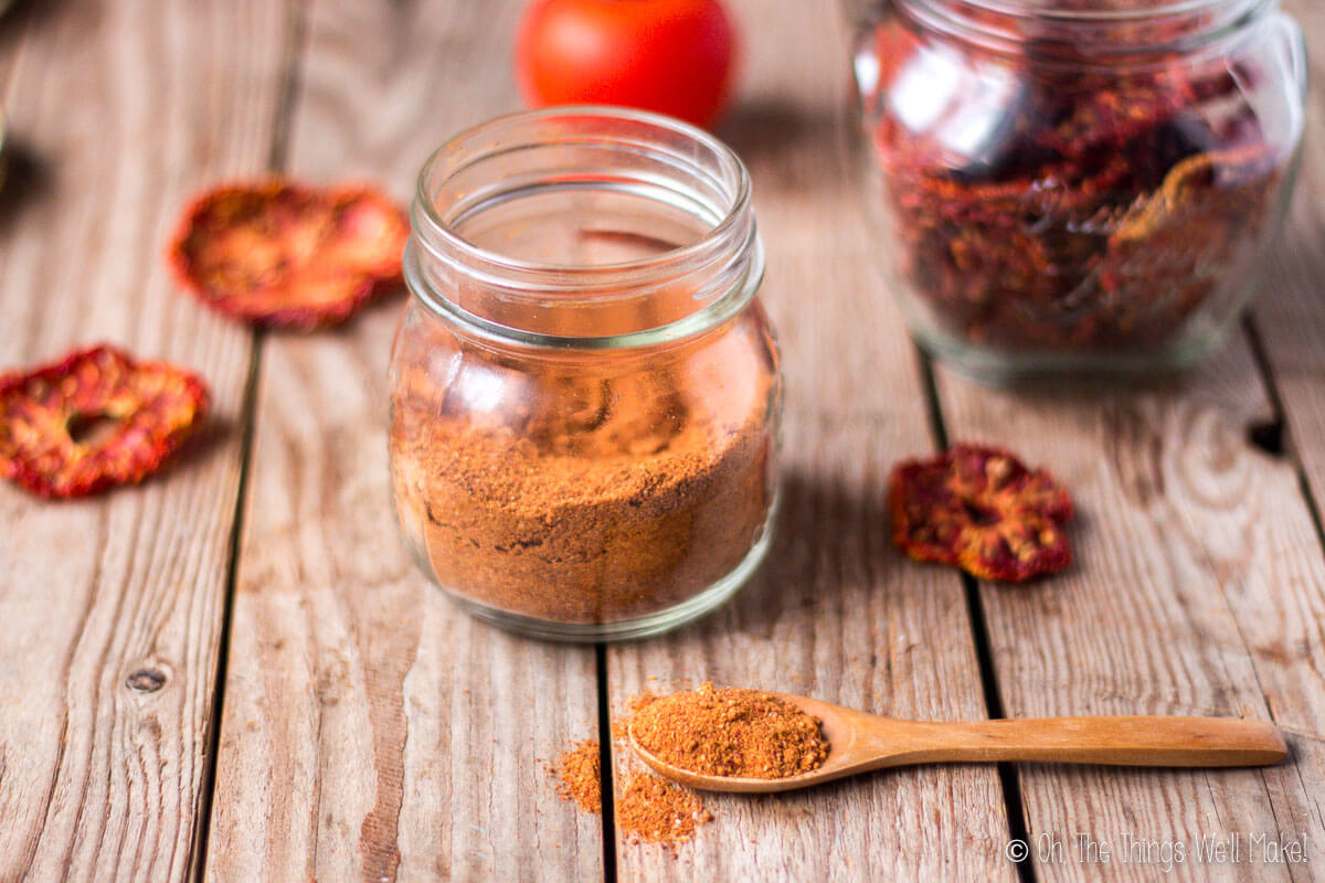 Tomato powder in a wooden spoon in front of a jar of tomato powder surrounded by dried tomato slices.