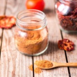 Tomato powder in a wooden spoon in front of a jar of tomato powder surrounded by dried tomato slices.