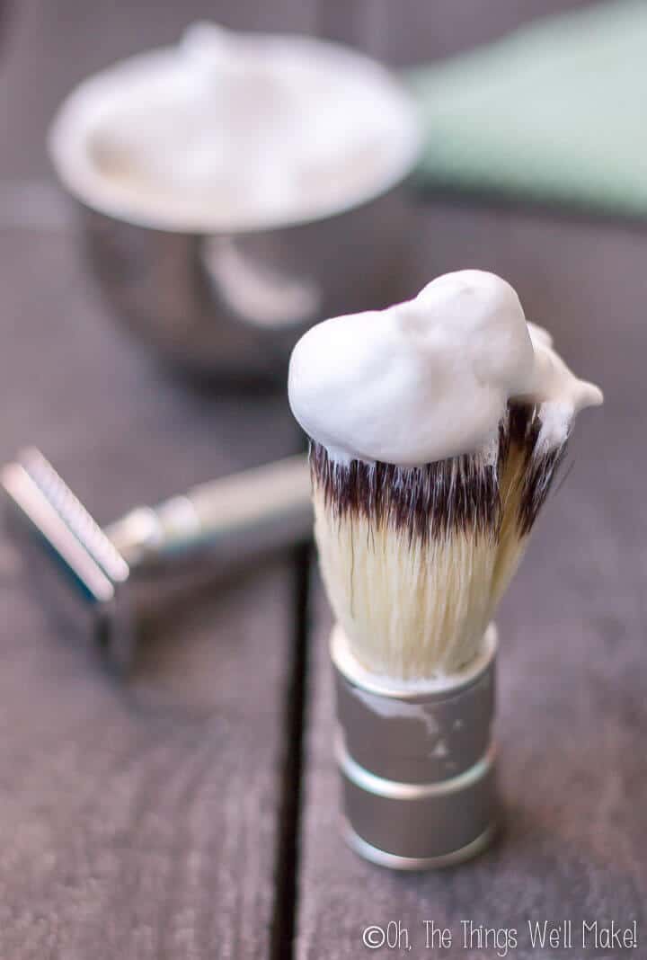 Closeup of a shaving brush with a thick shave soap lather on top. Behind, you can see the shave soap and a safety razor.