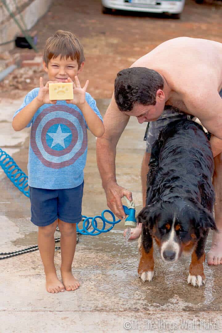 A boy holding a bar of soap next to his dad giving their dog a bath