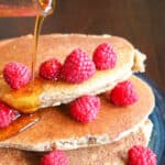 Almond pancakes stacked on a plate and covered with raspberries and maple syrup.