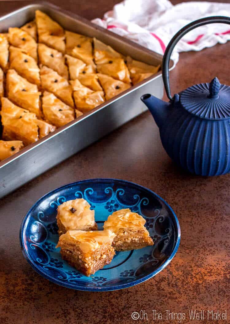 A few pieces of baklava on a plate in front of a pan full of baklava