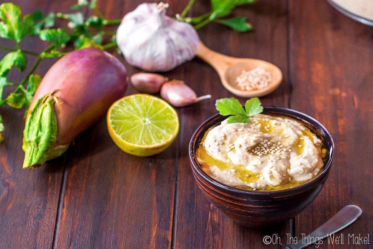 small bowl of baba ganoush in front of an eggplant, garlic head, and lemon half