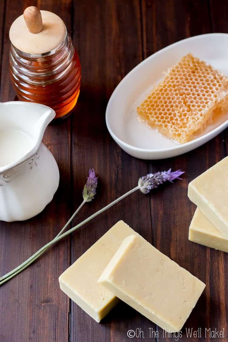 Bars of goat milk soap on a dark wood background next to a jar of honey, a honeycomb, some milk, and a couple of lavender flowers.