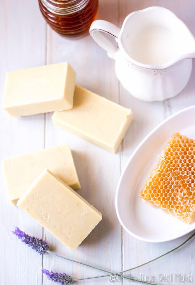 4 bars of goat milk soap with honey by a honeycomb and a pitcher of goat milk