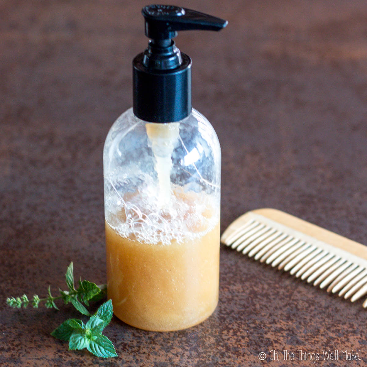 A bottle of shampoo in a pump bottle next to a wooden comb and a sprig of mint.