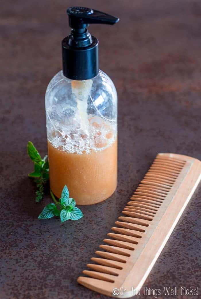 A homemade peppermint clarifying shampoo in a pump bottle next to peppermint sprigs and a wooden comb