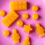 Bright orange homemade gummies in fun shapes, including LEGO bricks, LEGO figures, stars, and circles, arranged on a pink background.