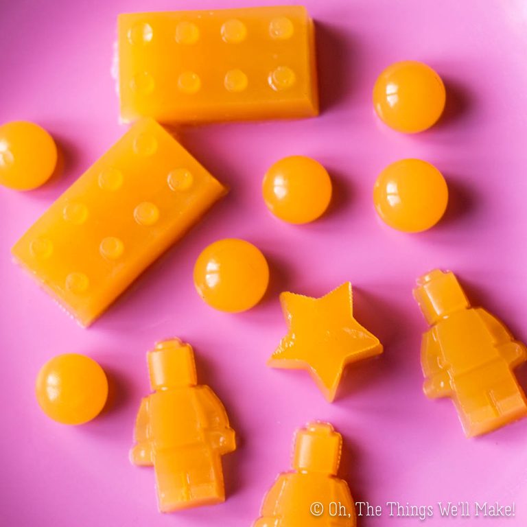 Bright orange homemade gummies in fun shapes, including LEGO bricks, LEGO figures, stars, and circles, arranged on a pink background.