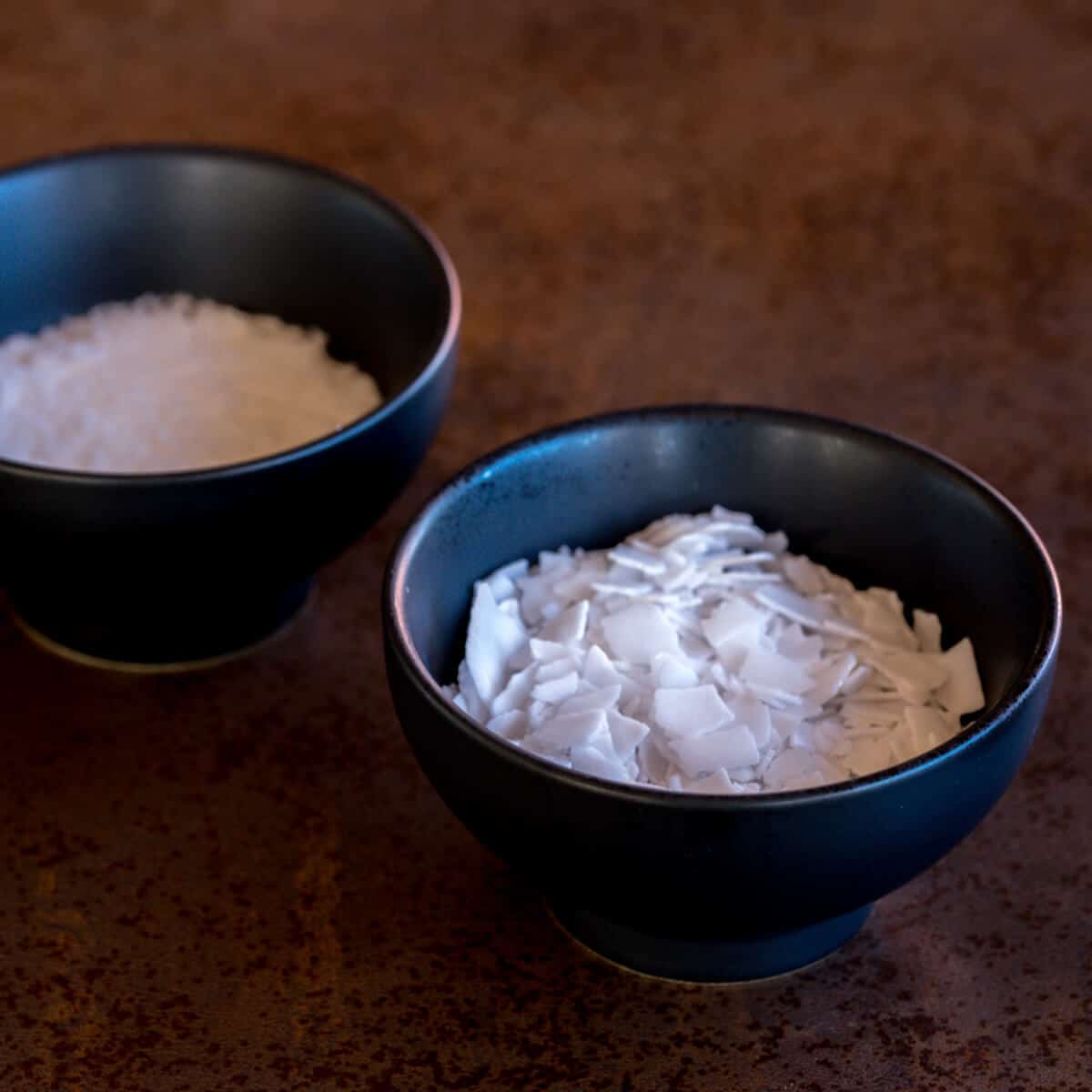 Overhead view of two bowls of lye, one with sodium hydroxide beads and another with potassium hydroxide flakes.