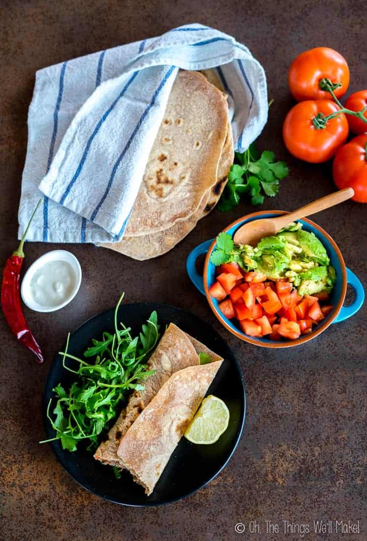 A wrapped chipotle shredded beef taco in front of homemade flour tortillas and other veggie sides and toppings.