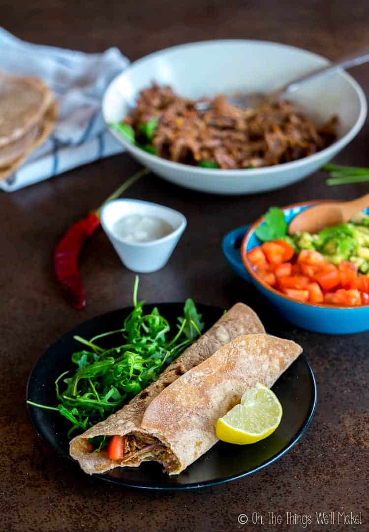 A wrapped chipotle shredded beef taco with a bite out of it. in front of a plate of shredded beef and some veggies and sides.