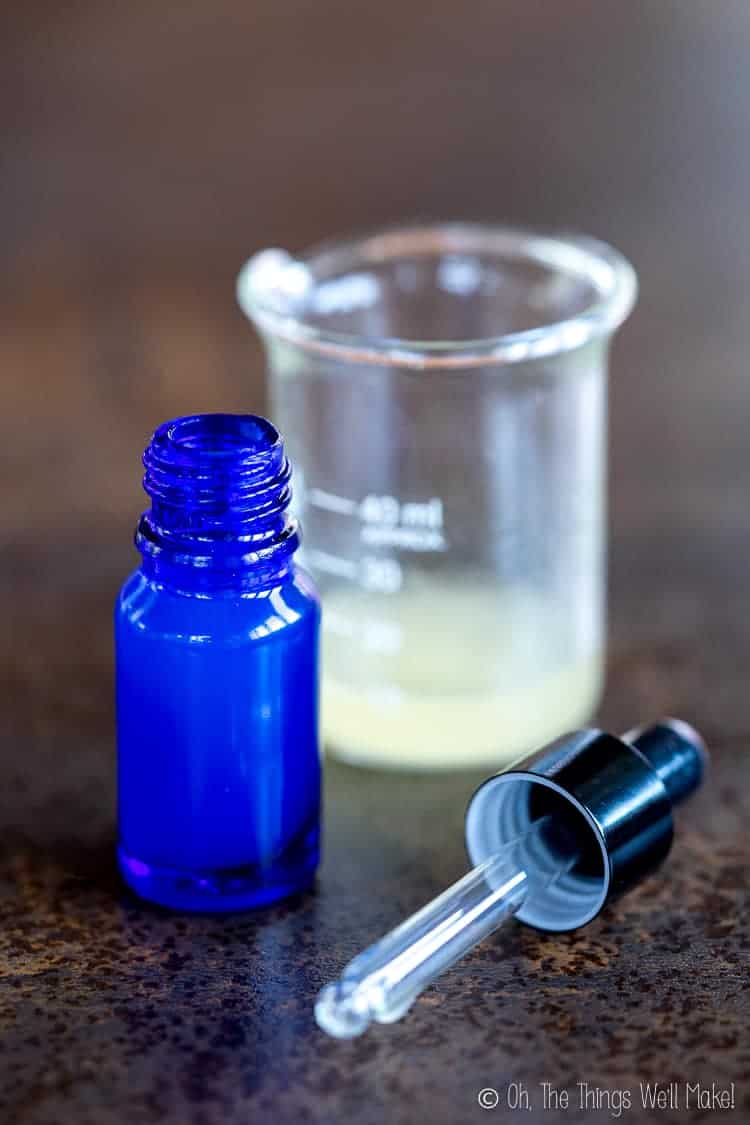 A beaker filled with a homemade hyaluronic acid serum, next to a blue glass bottle and glass dropper top.