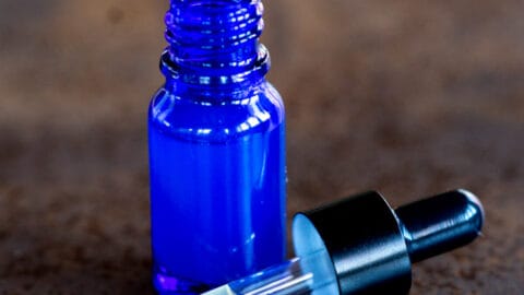A small glass blue bottle filled with a hyaluronic acid serum. The glass pipette is out and in front of the bottle, showing some of the serum inside it.