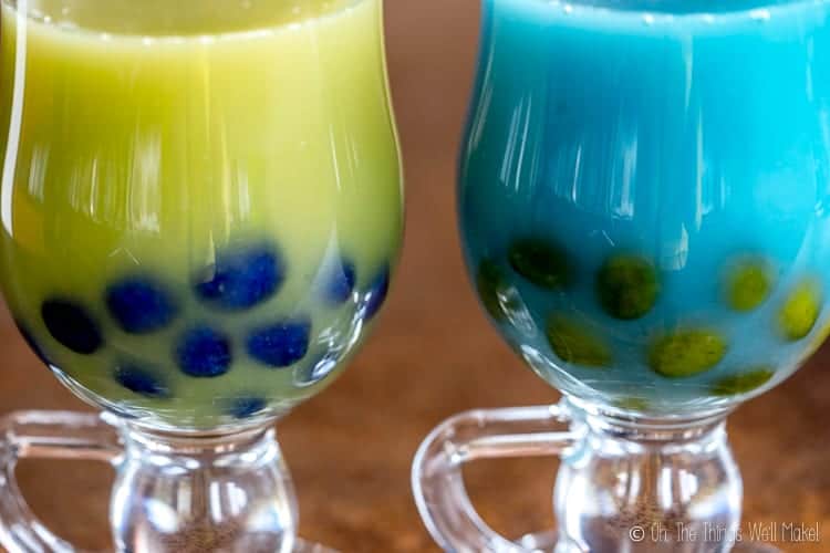 closeup of two bubble teas: one green with blue boba and one blue with green boba.