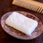 A homemade shampoo bar in a stone soap dish next to a wooden comb