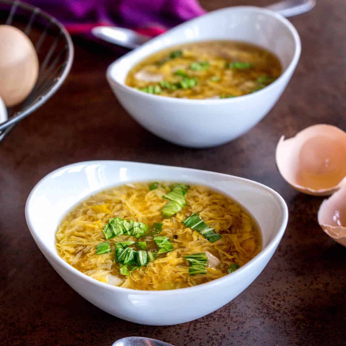 two bowls of egg drop soup with a basket of fresh eggs and open eggshells in the background.