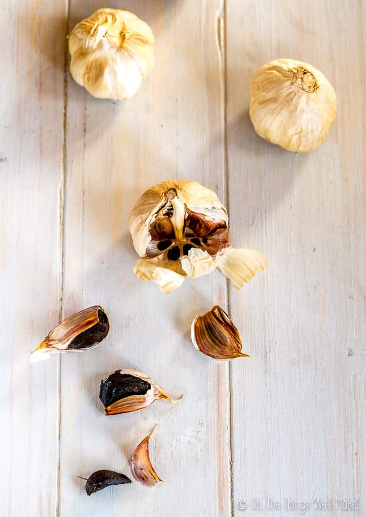 overhead view of several heads of black garlic, one opened exposing the black cloves, several of which have been opened to show their dark interior.