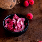 A bowl of raspberry seasoned coconut chips next to fresh raspberries and a coconut