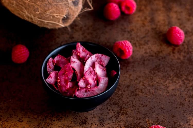 A bowl of raspberry seasoned coconut chips next to fresh raspberries and a coconut