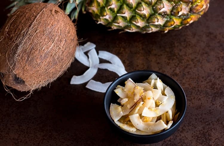 Overhead view of a bowl of piña colada seasoned coconut chips in front of a pineapple, coconut chips, and a fresh coconut.