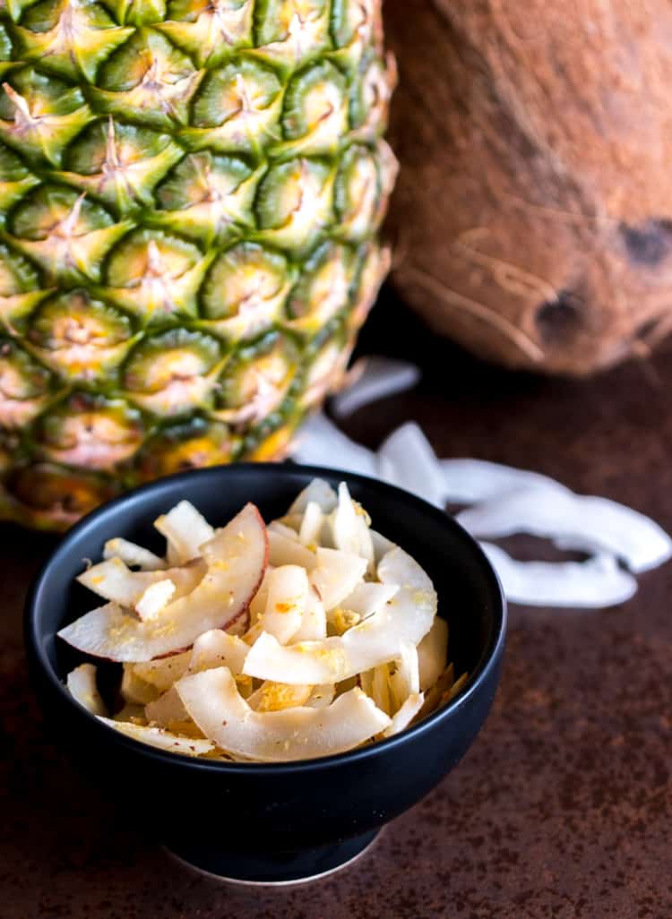 Piña colada seasoned coconut chips in a bowl in front of a pineapple and a coconut.