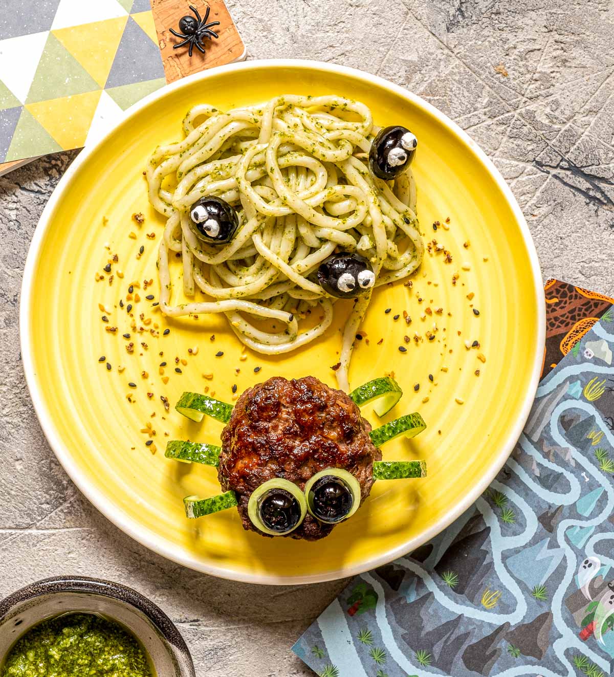 Overhead view of a plate of spaghetti with pesto sauce (a nest) covered with olives decorated with eyes next to a meatball "spider" with cucumber peel legs