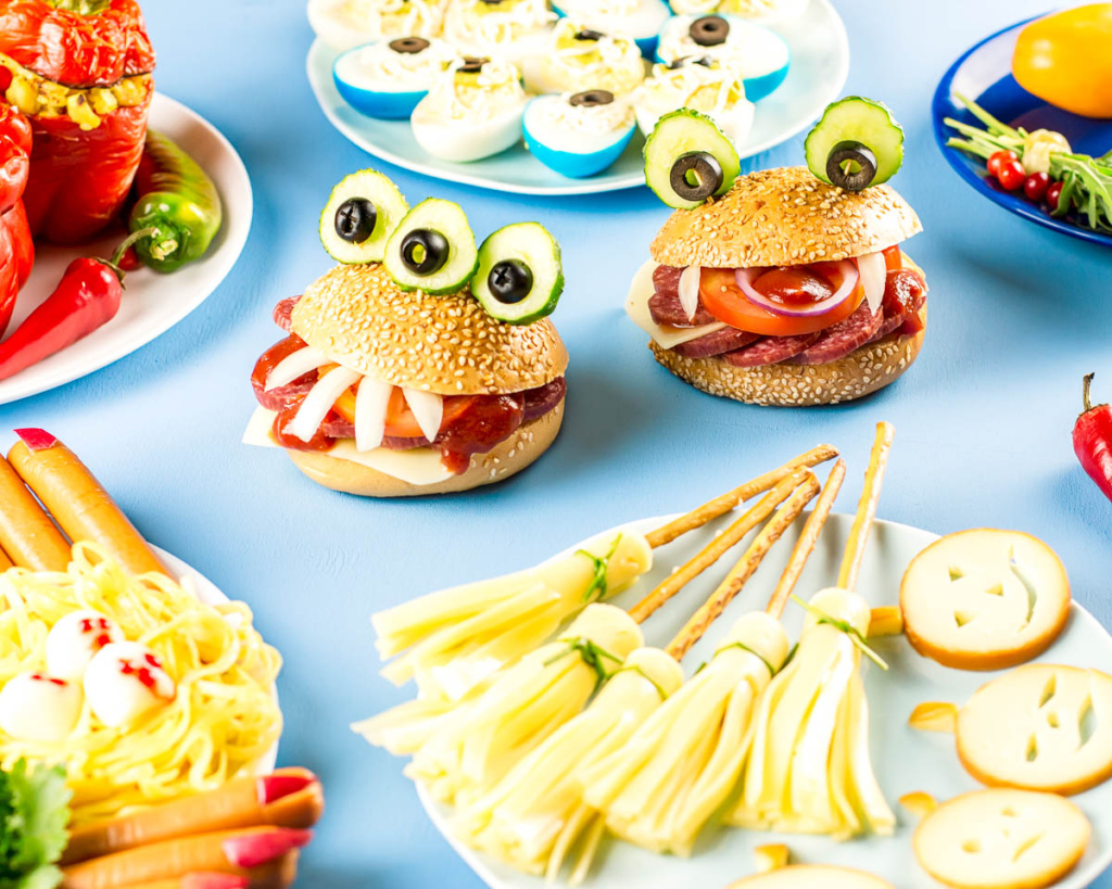 monster hamburgers next to a plate of pretzel and cheese broomsticks, frankfurt witche's fingers, and other Halloween foods.