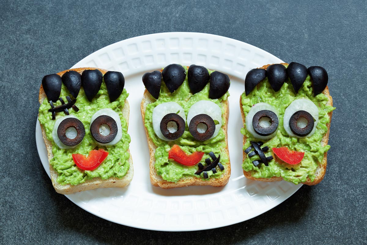 3 open-faced avocado sandwiches on a plate with Frankenstein faces made from black olives and red bell peppers. 