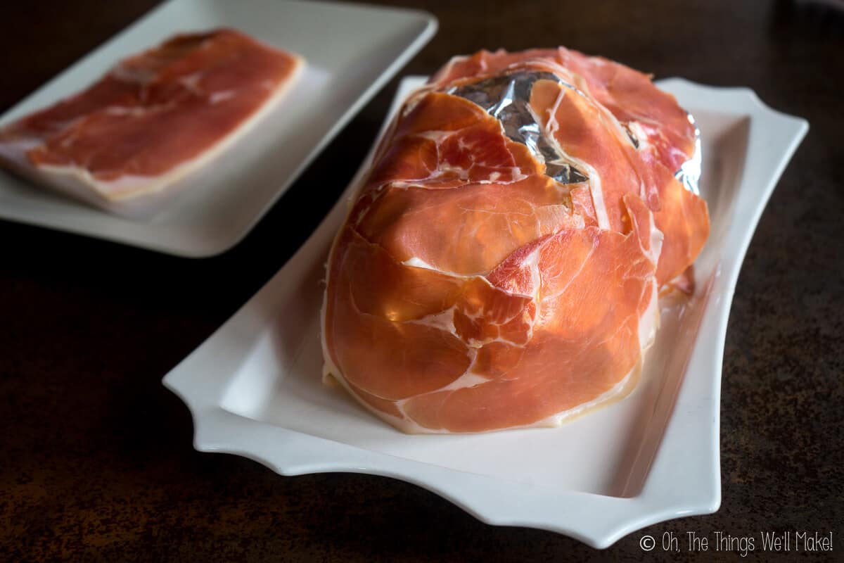 The foil covered face with more slices of prosciutto ham on it, on a white platter with a plate full of prosciutto ham on the left side.