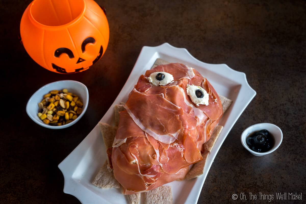 Overhead view of a creepy prosciutto face on a white plate beside a small bowl of nuts and a plastic Jack-o-Lantern.