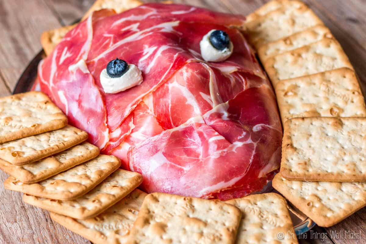 Prosciutto face with mozzarella cheese ball eyes and blueberry pupils on a platter surrounded by crackers