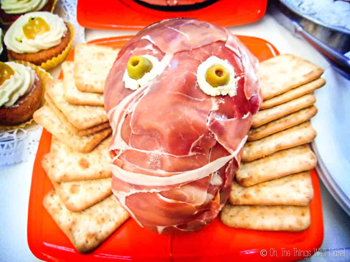 A prosciutto ham face with cream cheese eyeballs and olive pupils, surrounded by square biscuits.